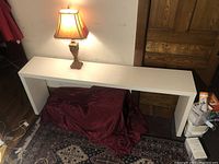 White Parsons style console table with wheels placed against wall with a lamp nearby. Surface flaw visible, table uncovered.