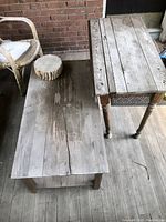 Photo of the low rectangular coffee table with wide wooden slats showing weathering and a round tree stump seat on top, placed on a porch wooden floor beside wicker chair.