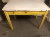 Full view of vintage utility table showing white enamel metal top and yellow painted wooden base with drawer and wooden knob.
