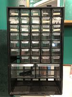 Front view of black plastic workshop organizer cabinet showing multiple clear drawers arranged in rows, some with labels, missing one small drawer and bottom larger drawer.