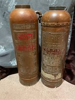 Two tall vintage brass fire extinguishers side by side, showing embossed labels and weathered patina.