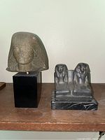 Photo depicting two Egyptian artifacts replicas on display: a bust of an Egyptian figure mounted on a tall black stand next to a smaller twin-figure statue on a black base.