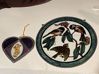 Two stained glass pieces displayed on a white surface: on the left, a small heart-shaped piece with purple and clear glass, a yellow floral inclusion, and metal frame; on the right, a larger oval piece with birds and leaves design and blue-green border.