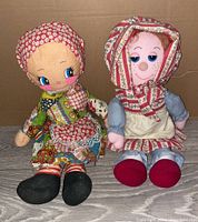 Two vintage Raggedy Ann style dolls seated side by side on a wooden surface against a cardboard background. One doll has a red floral bonnet and colorful patchwork dress, the other has a red and white striped bonnet and blue dress with apron.