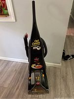 Full view of black Bissell upright carpet cleaner showing hose and side nozzle attachment, labels stating it cleans all leading brands.