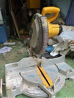 Side view of the DeWalt miter saw showing yellow handle and blade guard with DeWalt logo, resting on a green surface with visible dirt and debris.