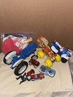 Photo of assorted vintage toys including a blue metal tractor, a wooden prancing pony pull toy with yellow wheels, several small toy vehicles, a handheld electronic game, and two red tennis paddles with balls in a plastic bag.