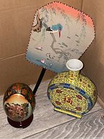 Photo showing a decorated large painted egg on wooden stand, yellow oriental-style ceramic bottle, and silk fan with sea bird design.