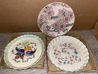 Three ceramic plates: two pie trays and one decorative plate shown together in cardboard boxes on floor.