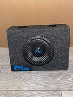 Front angled view of Ultimate Monitor Speaker in grey carpeted box with metal grille and blue logo.