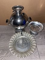 Front view of vintage metal samovar with two trays displayed beside it on a wooden floor in front of cardboard background.