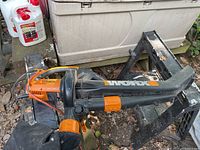 Worx electric leaf blower vacuum placed on black support structure outdoors among leaves and dirt, showcasing side and top view of the tool.