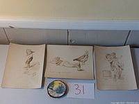 Three sepia lithograph prints and one round painted wood plaque laid out together on white surface with lot number card 31