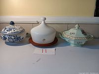 Three ceramic serving items side by side on shelf: a lidded blue and white soup tureen, a white milk glass bell-shaped piece on wooden base, and a green covered serving dish with floral patterns