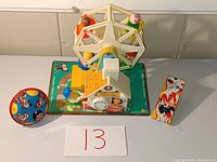 Photo showing the vintage Fisher Price metal toy carousel with four seated figures in orange, yellow, blue, and green, with a colorful base featuring a house and Ferris wheel graphics. Two metal clown toys with circus designs sit beside it.