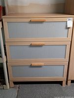 Front view showing dresser with three drawers having translucent plastic fronts and wooden handles. Light wood finish with minor wear marks.