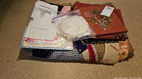 Lot contents showing folded embroidered napkins in plastic bag, doilies in a plastic zip bag, framed embroider floral art piece, and hand stitched patchwork quilt beneath on carpeted floor.