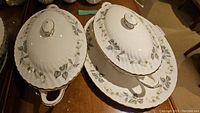 Two covered serving bowls and one covered serving platter all with leaf and vine pattern and gold trim on a wood surface.