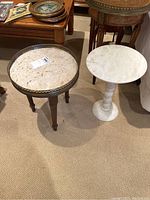 Two side tables placed on carpeted floor; one with wood legs, stone top and brass rim, the other a small white marble pedestal side table.