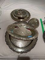 Four silverplate serving pieces displayed on a white cloth, showing a large round tray, an oval tray, a covered oval serving dish, and a small round bowl, all with embossed designs and tarnishing.