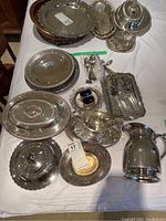 Full overhead view of assortment including trays, spoons, jugs, covered serving dish, salt and pepper shakers laid out on white surface showing tarnishing and reflective surfaces.