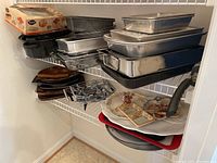 Shelves showing stacked metal baking pans, roasting pans, and assorted ceramic serving plates and trays.