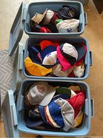 Three plastic storage bins filled with assorted ball caps in multiple colors, with lids included.