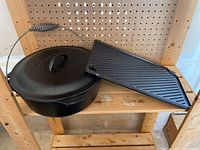 Cast iron Dutch oven with lid and griddle placed on wooden shelf, showing their prepared condition and size.