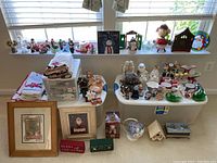 Wide view of the assortment of Christmas figurines, towels stacked and folded, framed Christmas artwork, and ceramic decorative items on a table and in front of a window