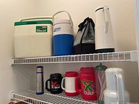 Shelving with various coolers and thermoses, showing Oscar cooler, Coleman jug, soft cooler bag, and assorted insulated mugs