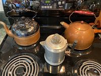 Photo showing all three kettles on a stove top: a copper and black kettle, copper kettle with wooden handle, and aluminum kettle with glass lid.