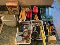 Wide view showing assortment of kitchen utensils and tools organized in plastic bins including whisks, bamboo skewers, BBQ utensils, measuring spoons, cookie cutters, and pepper grinders