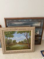 Photo showing assorted large framed paintings leaning against wall. Foreground includes a framed landscape painting with green trees and a decorative beige frame.
