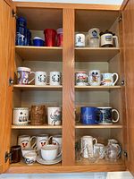 Wooden cabinet shelves containing a variety of mugs and glassware including collectible and novelty mugs, ceramic and glass.