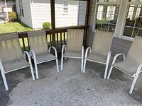 Five white metal framed outdoor chairs with beige mesh seats and backs arranged side by side on a concrete patio.