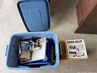 Open plastic storage bin showing assorted painting tools including paint trays, brushes, rollers, and the Wagner Power Roller box