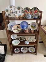 Full view of wooden shelf displaying various decorative plates of different sizes and designs, some with stands, plus porcelain cups and small boxes.