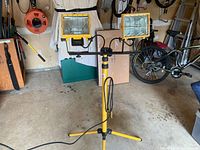 Yellow dual flood lights mounted on a tripod stand in a garage setting with one bulb unlit.