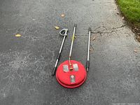 Red circular surface cleaner pressure washer attachment with three metal extension rods and spray gun handle, placed on driveway.