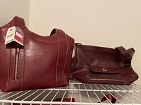 Two large burgundy purses on a white shelf, one with a zipper and tag attached, other with front pocket and clasp