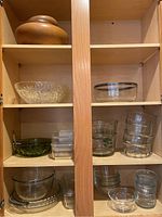 Photo showing wooden bowls on top shelf, various clear glass bowls, glass measuring cups with floral or fruit designs, small clear glasses and other glassware.