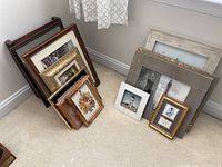 Photo showing assortment of framed small wall art leaning against a wall on carpet, various frame styles and sizes visible.