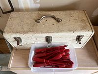 Closed white metal tool box sitting on a table with a clear plastic bin of red-handled screwdrivers in front.