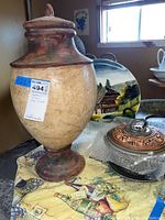 Large urn-shaped clay jar with lid, clay jar placed on a decorative cloth with fruit and wine motifs, a large decorative ceramic plate with chalet scene in background, copper roaster with lid wrapped in bubble wrap.