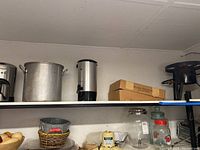 Shelf containing large metal pot, silver coffee urn, and boxed manual on upper shelf; baskets, jars, and metal containers on lower shelf