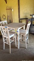 Wooden high table with three matching wooden high chairs in natural wood finish, photographed in a rustic indoor environment with pool cues and other furniture visible.