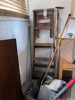 Full view of wooden ladder, garden hoe with wooden handle and metal blade, broom, mop, and metal mop bucket on caster wheels with mop wringer attached.