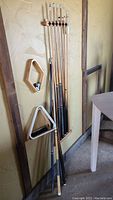 Photo showing the pool cue sticks hung vertically on the wall rack along with the two different style pool racks mounted on the wall close to the cues.