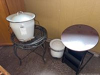 Photo showing large white porcelain container on black metal round table, small porcelain container on floor, black metal stool and magazine rack nearby.