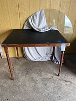 Photo showing a handcrafted wooden table with black surface and foldable legs. A circular glass tabletop is positioned behind the table.